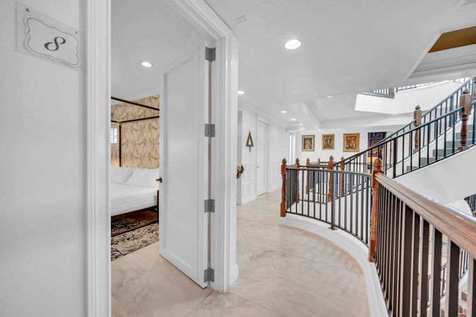 Bright second-floor hallway and curved wood-and-iron staircase in a residential interior, with recessed lighting, framed artwork on the landing, and an open bedroom door showing patterned wallpaper and a white bed.