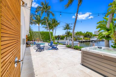 Tropical waterfront patio with hot tub and outdoor shower, pool with cascading waterfall, blue Adirondack chairs around a fire pit, string lights and palm trees overlooking a boat dock and canal.