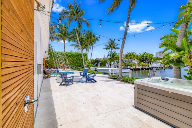 Tropical waterfront patio with hot tub and outdoor shower, pool with cascading waterfall, blue Adirondack chairs around a fire pit, string lights and palm trees overlooking a boat dock and canal.