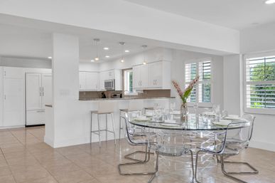 Bright modern open-plan kitchen and dining area with white cabinetry, granite breakfast bar and stools, glass round table with clear acrylic chairs, tile floor, plantation shutters and a vase of flowers.
