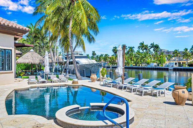 Tropical waterfront pool and spa overlooking a marina with a docked yacht, swaying palm trees, a row of sun loungers and umbrellas under a bright blue sky