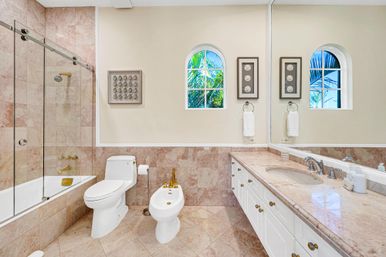 Bright luxury marble bathroom with glass-enclosed bathtub, gold fixtures, toilet and bidet, long vanity with double sink and large mirror, and arched windows framing tropical palm views.