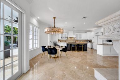 Bright open-plan luxury kitchen and dining area with glossy beige marble floors, white cabinetry, large black island with gold-accent bar stools, round dining table surrounded by navy velvet chairs under a crystal chandelier, and French doors opening to a tropical patio.
