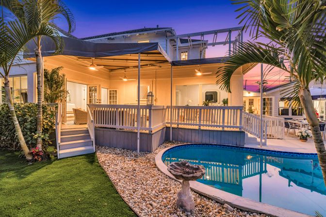 Sunset-lit tropical backyard with curved swimming pool, pebble border and decorative birdbath, raised covered wooden deck with warm lights and rattan seating, palm trees and manicured lawn for a cozy coastal patio scene.
