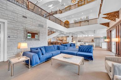 Bright luxury open-concept living room with a large blue sectional, stone accent wall, mezzanine balconies, vaulted ceiling, and open kitchen in the background.