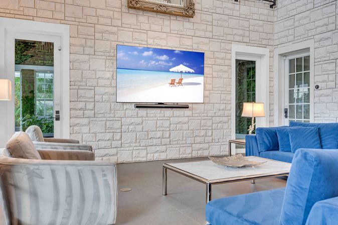 Relaxing coastal-inspired living room with white stone accent wall, wall-mounted TV showing a beach scene, blue velvet sofa, gray armchairs, marble-top coffee table and table lamps.