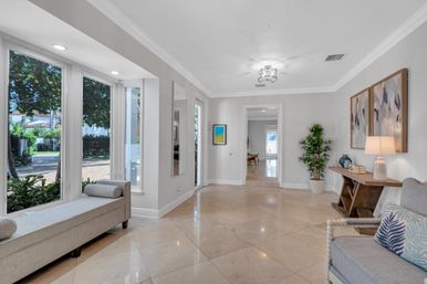 Sunlit, inviting foyer with floor-to-ceiling windows overlooking a tree-lined street, beige marble tile floor, window bench, console table with lamp, abstract wall art and potted plant.