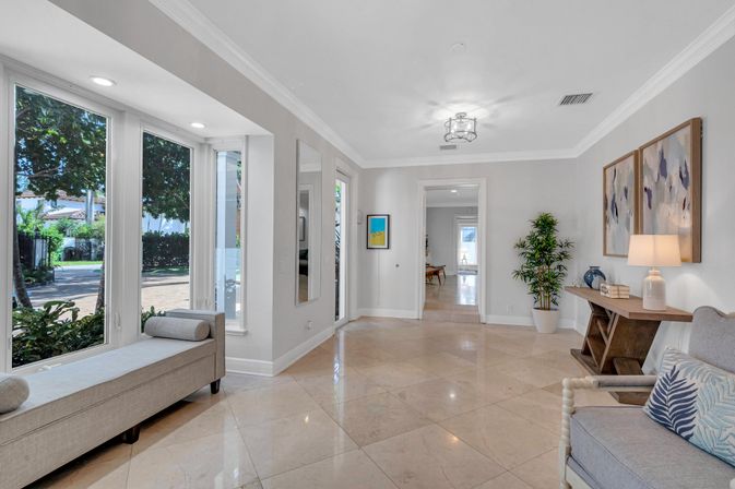 Sunlit, inviting foyer with floor-to-ceiling windows overlooking a tree-lined street, beige marble tile floor, window bench, console table with lamp, abstract wall art and potted plant.