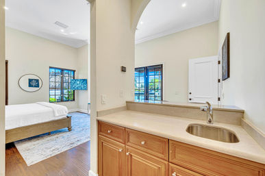 Bright, sunlit bedroom suite with white bed and coastal wall art seen through an arched pass-through to a marble-topped wet bar with sink and wood cabinets, large windows and glass doors opening to a sunny patio with palm trees.