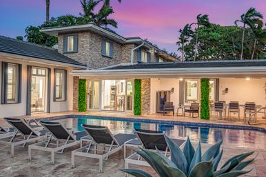 Luxury modern two-story tropical home with stone facade and open sliding doors onto a tiled patio, evening pool bordered by a row of lounge chairs, covered outdoor dining and seating, palm trees and pink-purple sunset sky.