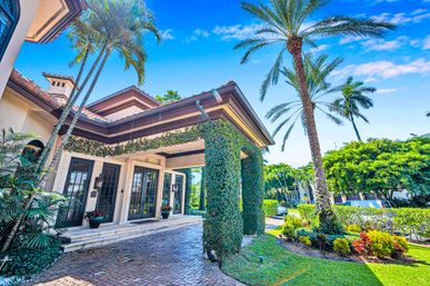 Sunlit luxury Mediterranean-style home entrance with ivy-wrapped columns, glass French doors, palm trees and manicured tropical landscaping.