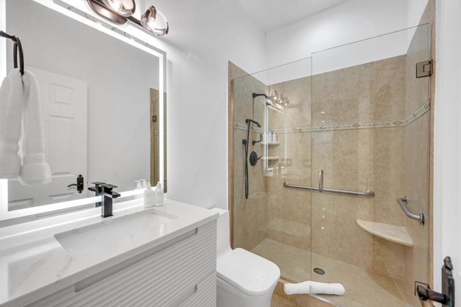 Modern white bathroom with LED-lit vanity mirror, black faucet and quartz countertop; glass-enclosed beige-tiled walk-in shower with bench, handheld showerhead and stainless grab bars.