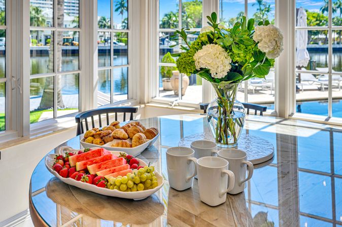 Sunny waterfront breakfast table in a tropical canal-view dining nook with platters of watermelon, strawberries, grapes, pastries, four coffee mugs and a hydrangea bouquet visible through large windows