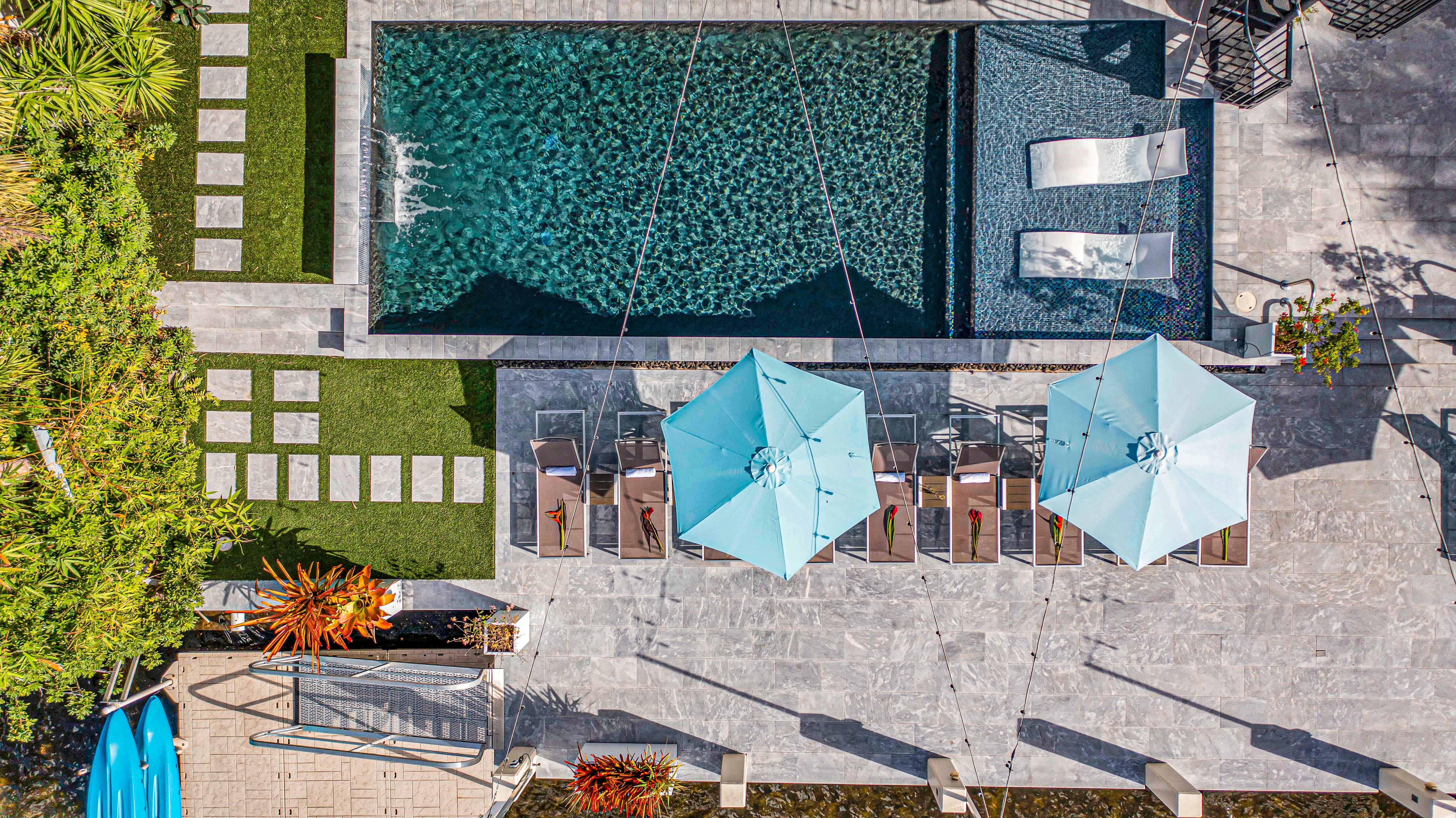 Aerial view of a modern backyard pool: rectangular turquoise pool with two in-water loungers, row of sunbeds under blue umbrellas on a stone patio, stepping-stone lawn, tropical plants and kayaks