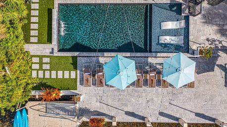Aerial view of a modern backyard pool: rectangular turquoise pool with two in-water loungers, row of sunbeds under blue umbrellas on a stone patio, stepping-stone lawn, tropical plants and kayaks