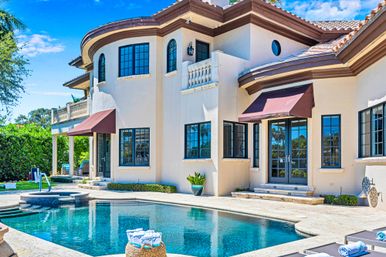 Sunny Mediterranean-style two-story home with terracotta roof, arched windows, brown awnings and a sparkling backyard pool with stone deck and palm trees