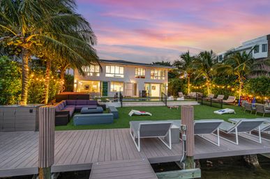 Tropical waterfront sunset scene: modern two‑story house with lit palm trees, dock and lounge chairs, outdoor sectional seating, grassy lawn with cornhole boards and a pool