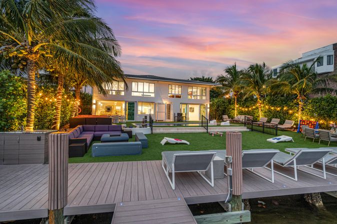 Tropical waterfront sunset scene: modern two‑story house with lit palm trees, dock and lounge chairs, outdoor sectional seating, grassy lawn with cornhole boards and a pool
