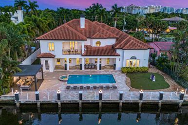 Aerial view of a Mediterranean-style waterfront villa with terracotta roof, lit windows, backyard pool and spa, row of loungers, covered patio seating, private dock on a calm canal, palm trees and purple sunset sky.
