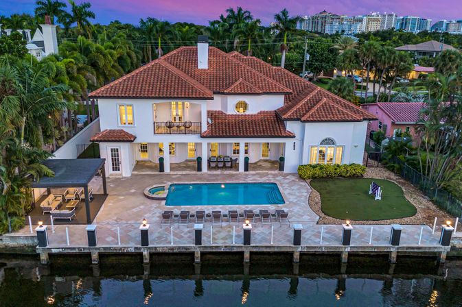 Aerial view of a Mediterranean-style waterfront villa with terracotta roof, lit windows, backyard pool and spa, row of loungers, covered patio seating, private dock on a calm canal, palm trees and purple sunset sky.
