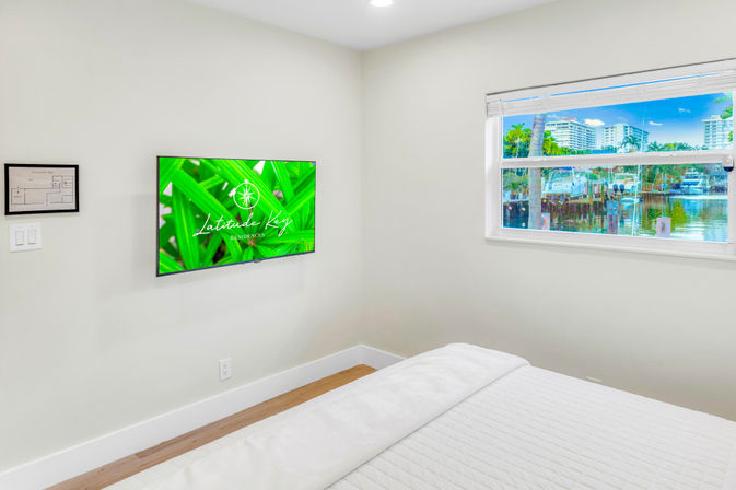 Sunny modern coastal bedroom with wall-mounted TV showing green foliage, white quilted bed, and a window view of a marina with boats, palm trees, and waterfront buildings.