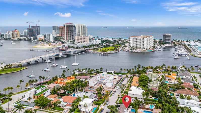 Aerial view of a sunny tropical waterfront neighborhood with palm-lined streets, pools and luxury homes, a bridge to a marina full of sailboats and a high-rise ocean skyline with cargo ships on the horizon.