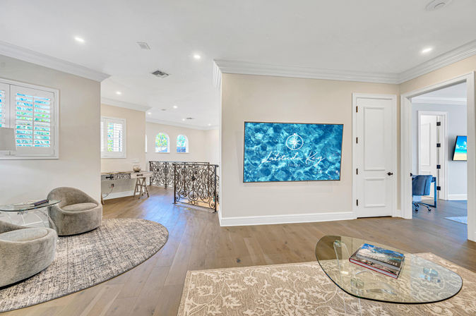 Bright coastal-style upstairs lounge with hardwood floors, round gray swivel chairs on a circular rug, glass coffee table with books, ornate wrought-iron railing, and a wall-mounted TV showing a blue ocean graphic