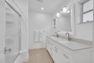 Bright modern master bathroom with double-sink white vanity, marble countertop, brushed-nickel faucets, large mirror with lights, frosted-glass shower door, white towels on a rack, and beige tile floor.