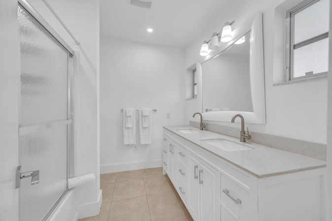 Bright modern master bathroom with double-sink white vanity, marble countertop, brushed-nickel faucets, large mirror with lights, frosted-glass shower door, white towels on a rack, and beige tile floor.