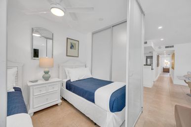 Bright, contemporary guest bedroom with two twin beds in navy quilted bedding, white nightstand and lamp, ceiling fan, sliding closet doors, and a hallway opening to an open-plan kitchen and dining area—coastal-inspired vacation rental style.
