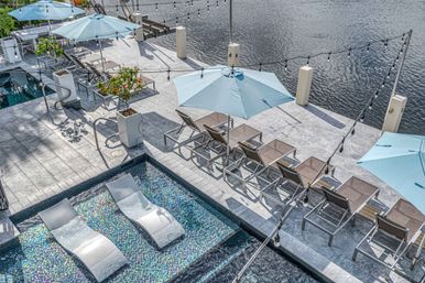 Aerial view of a waterfront resort pool deck with a mosaic-tiled sun shelf and two submerged lounge chairs, rows of gray sunbeds under pale blue umbrellas, potted plants, hanging string lights and calm water beyond.