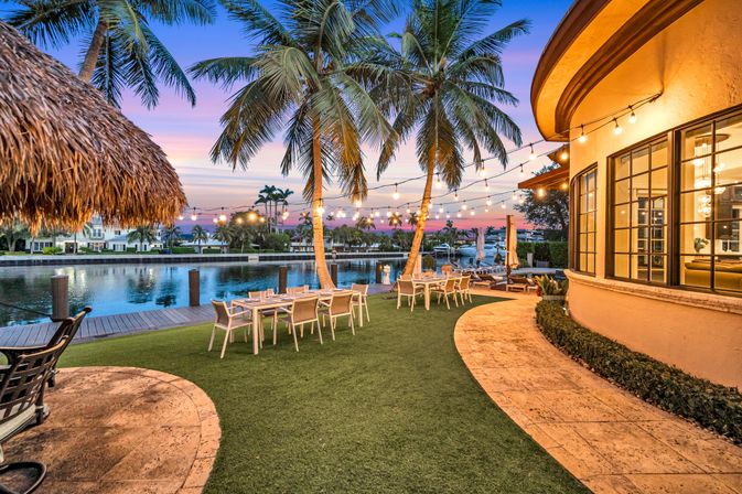 Tropical waterfront backyard at sunset with swaying palm trees, hanging string lights above outdoor dining tables, curved stone patio and dock along a calm canal.