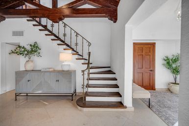 Airy Mediterranean-style foyer with floating dark-wood stairs and ornate wrought-iron railing, exposed wooden beams, cream tile floor, gray ribbed console table with lamp and potted greenery, and a wooden interior door.