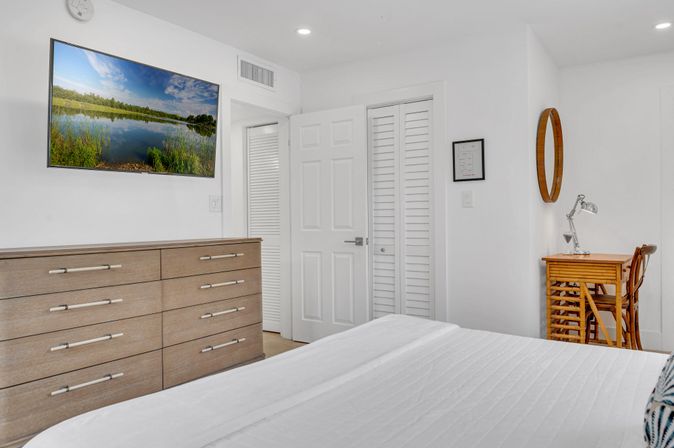 Bright modern bedroom with white walls and bedding, light-wood dresser, wall-mounted flat-screen TV showing a lake scene, louvered closet doors, and a small wooden desk with lamp and round mirror — clean, coastal-inspired decor.