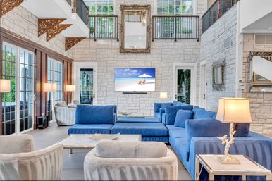 Bright luxury coastal living room with deep-blue sectional and cream armchairs, stone accent wall, wall-mounted TV showing a beach scene, floor-to-ceiling glass doors, high-ceiling indoor balcony and decorative mirrors.