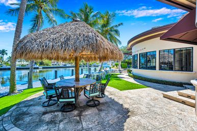 Sunny tropical canal-side patio with large thatched palapa over round dining table, cushioned chairs, palm trees and dock