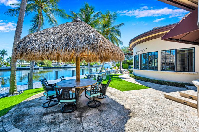 Sunny tropical canal-side patio with large thatched palapa over round dining table, cushioned chairs, palm trees and dock