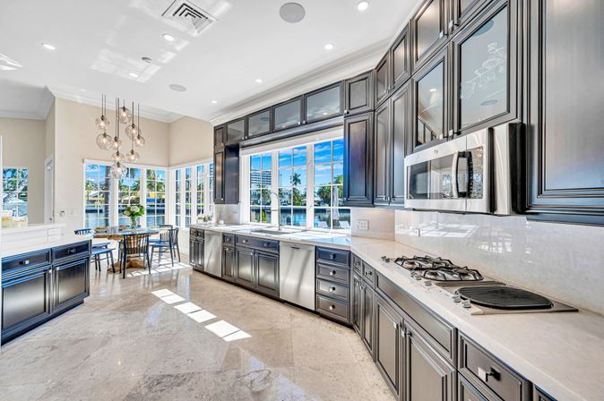 Sunlit luxury waterfront kitchen with dark cabinetry, marble floors and counters, stainless appliances and gas cooktop, plus a bright bay-window breakfast nook overlooking water and palm trees.