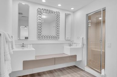 Bright contemporary white bathroom with double floating vanity and sinks, decorative linked-frame mirror, two narrow mirrors, glass-enclosed tiled shower with bench, wood-look floor and recessed lighting
