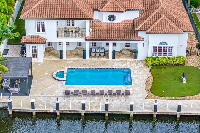 Aerial view of a Mediterranean-style waterfront home with red-tile roof, rectangular turquoise pool, row of lounge chairs, covered patio dining area, small putting green, and private dock.