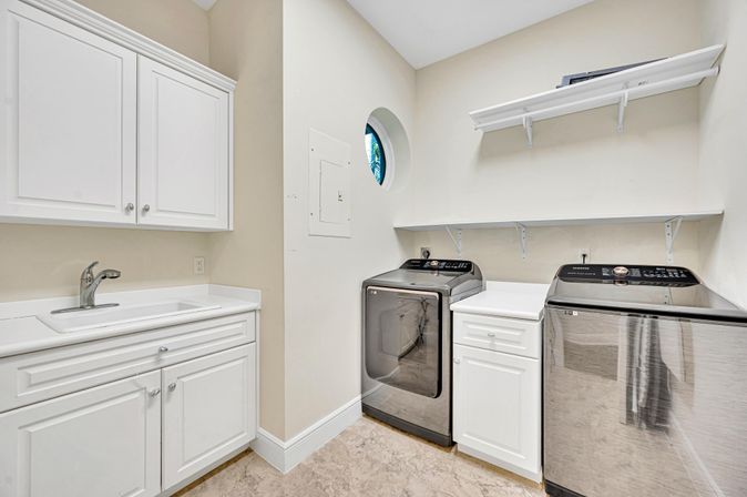 Bright modern laundry room with stainless steel front-load washer and dryer, white cabinets and utility sink, open wall shelves, round window and beige tile floor.