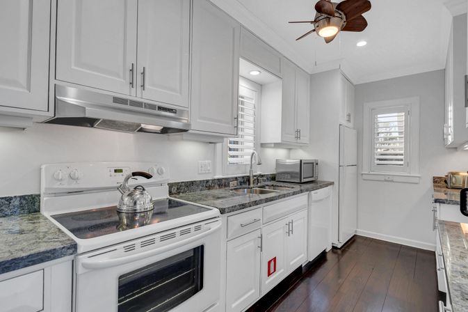 Bright modern white kitchen with shaker cabinets, granite countertops, stainless-steel kettle on an electric range, microwave and double sink, white fridge, dark hardwood floors and ceiling fan