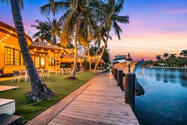 Tropical waterfront dock at sunset with palm trees, twinkling string lights, outdoor dining area and a moored yacht along a calm canal.
