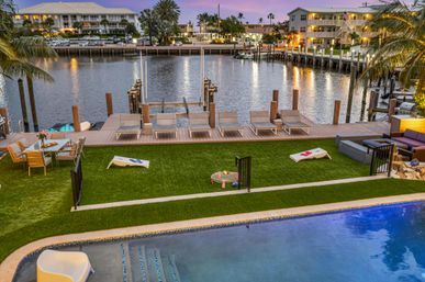 Waterfront canal backyard at sunset with pool, artificial-turf lawn, cornhole boards, dining area, lounge chairs on a wooden dock, palm trees and waterfront condos
