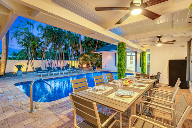 Tropical evening backyard with a covered patio and ceiling fans, a long outdoor dining table set for a meal, and a lit private swimming pool with lounge chairs and palm trees.