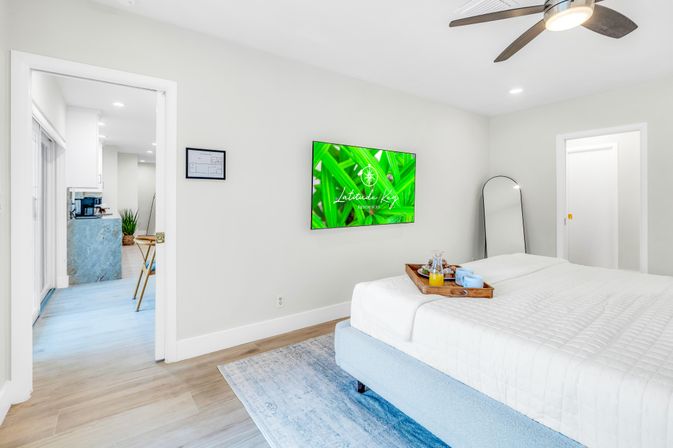 Bright modern bedroom with king bed and white quilt, wall-mounted TV showing green leaves, ceiling fan, full-length mirror, breakfast tray and open doorway to kitchen