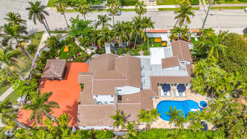 Aerial view of a luxury Florida coastal home on a corner lot with bright blue pool and spa, palm-lined tropical backyard, orange patio with tiki hut, lounge chairs, and lush landscaping.