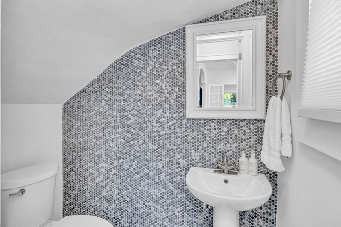 Cozy small bathroom/powder room with gray penny-tile mosaic accent wall, white pedestal sink, framed mirror, towel ring and toilet under a sloped ceiling