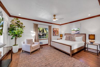 Bright, spacious bedroom with king bed, patterned rug, upholstered armchair, potted fiddle-leaf fig and palm trees visible through the window, warm wood trim and modern ceiling fan.