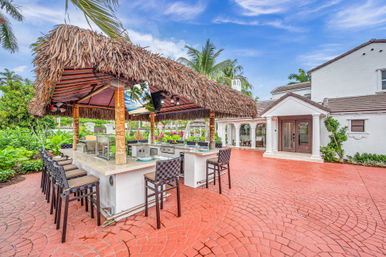 Sunny tropical tiki bar and outdoor kitchen with thatched roof, bar stools on a red brick patio beside a white Mediterranean-style luxury home, palm trees and blue sky overhead.